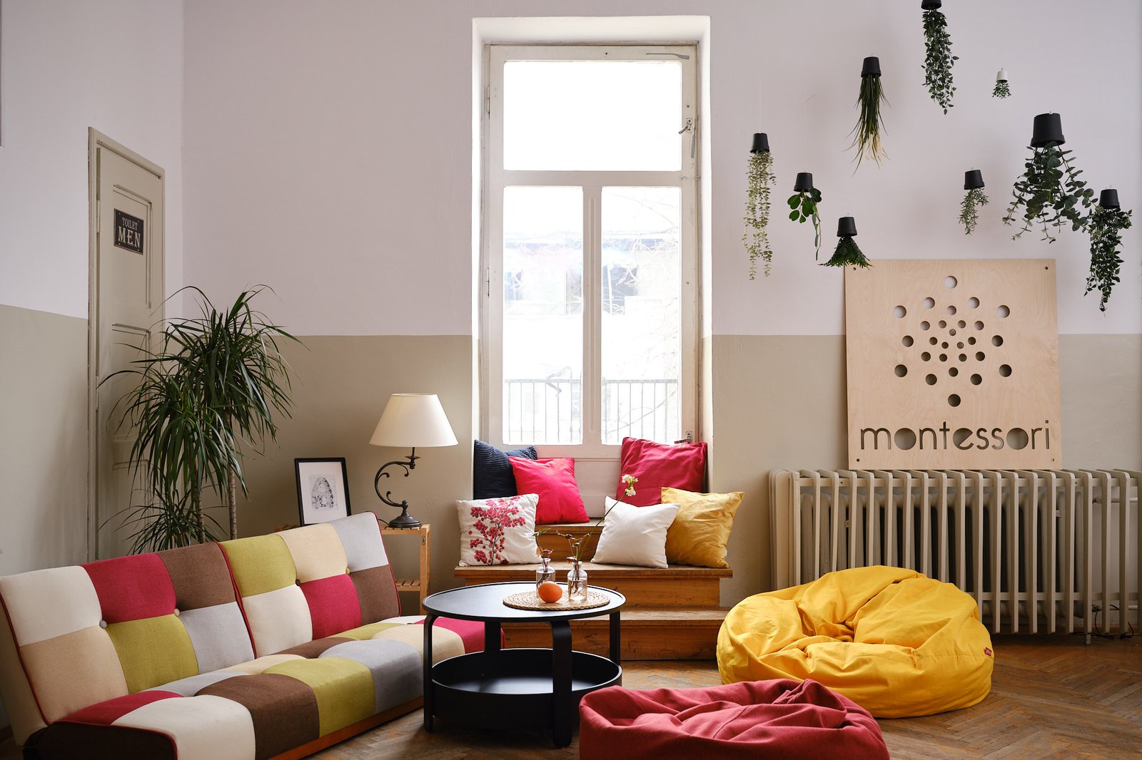 Przytulna strefa relaksu w liceum Montessori z kolorową sofą, pufami, poduszkami i roślinami wiszącymi przy dużym oknie